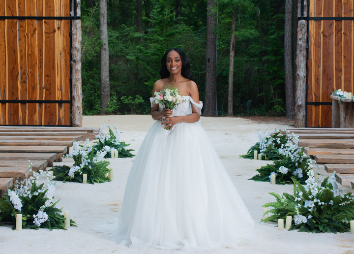 A woman in a white off-the-shoulder ball gown wedding dress stands on a sandy aisle holding a bouquet, framed by wooden doors, trees, and floral arrangements.